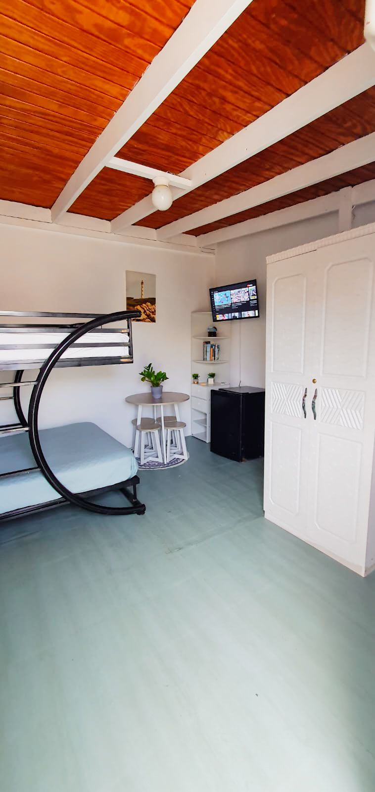 The cozy studio features a black metal bunk bed against a light-colored wall, with a mattress on the lower level. A small table with two stools is positioned nearby, and a compact refrigerator sits beside a white wardrobe. A television mounted on the wall provides entertainment.