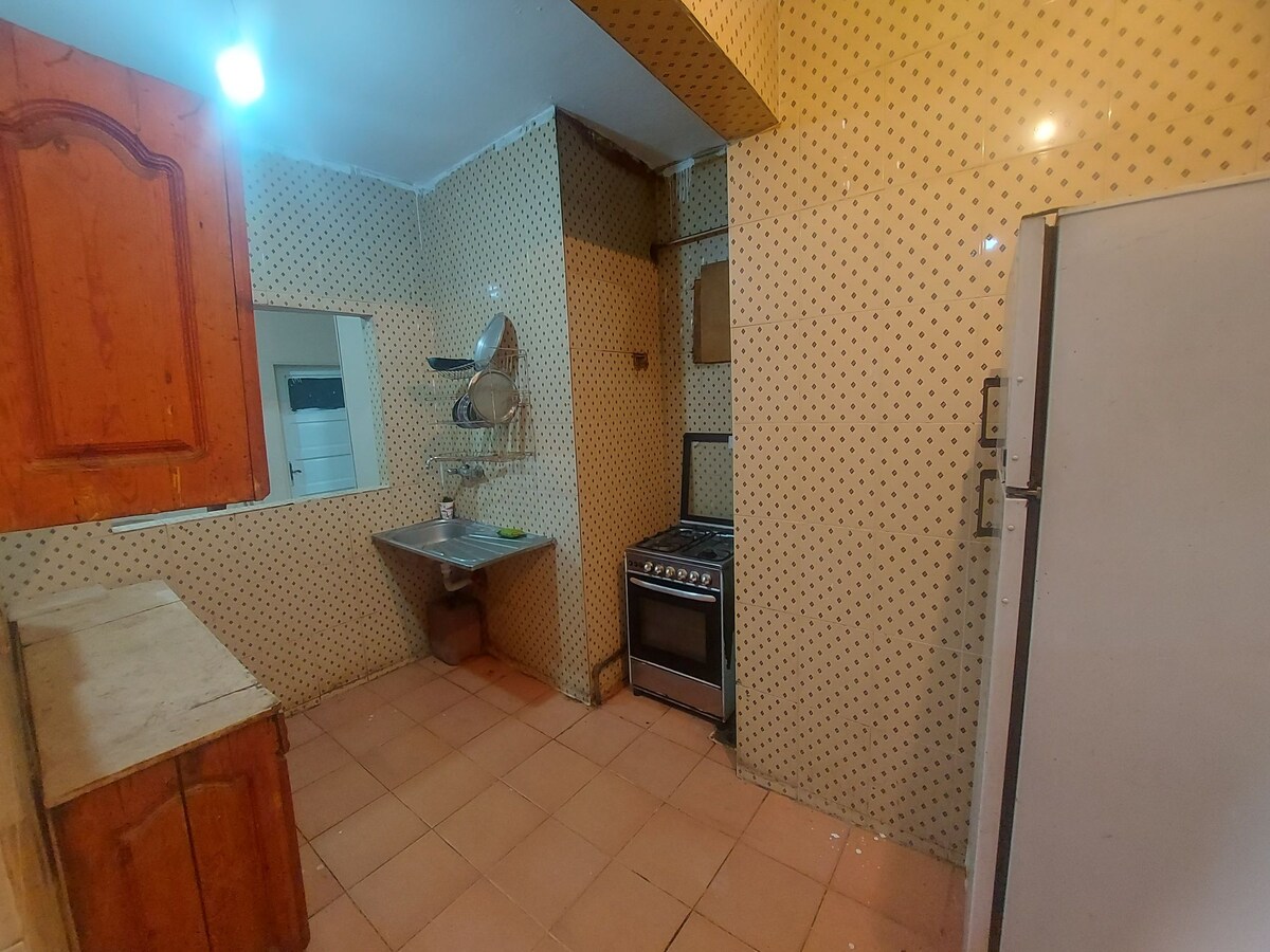 A kitchen space is presented with patterned walls and tiled flooring. Counter space includes wooden cabinets and a stainless steel sink. An oven and stove are visible alongside a refrigerator. Natural light enters through an open window, enhancing the room's functionality.