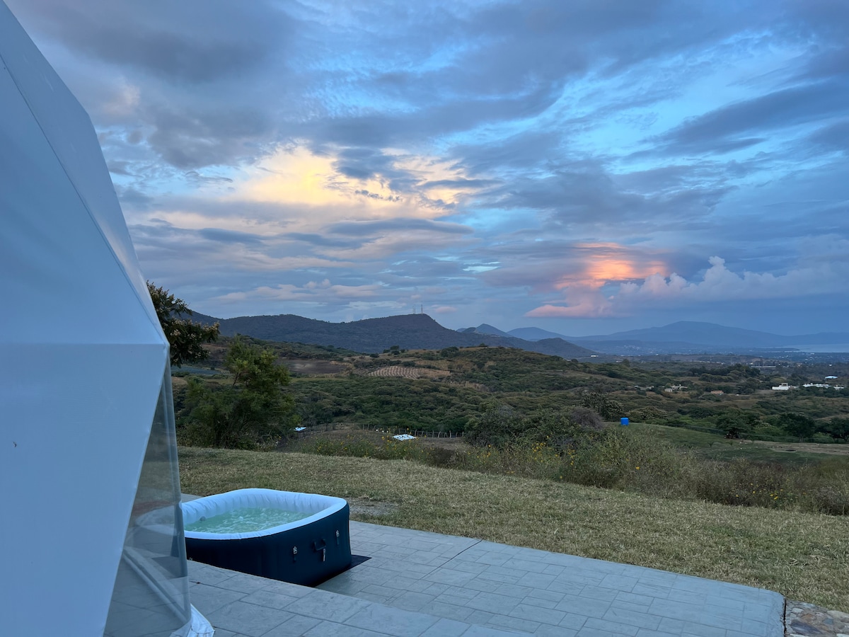 A clear view of the surrounding landscape is presented, showcasing rolling hills under a soft sky with pastel hues at sunset. A hot tub is positioned on the patio, inviting relaxation while enjoying the natural scenery.