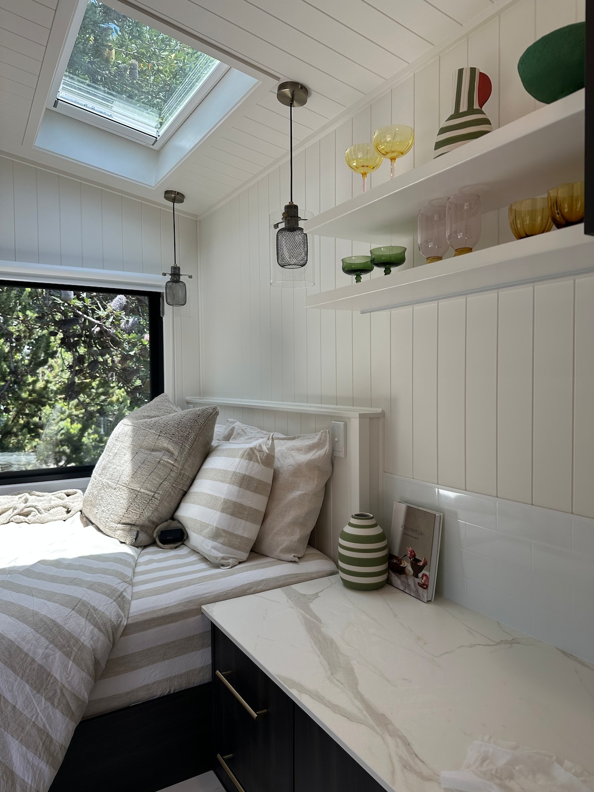 A cozy sleeping area features a spacious bed layered with striped blankets and decorative pillows. Above, a skylight offers natural light, while shelves display an array of colorful glassware, enhancing the inviting design. A bedside surface provides additional space for personal items.