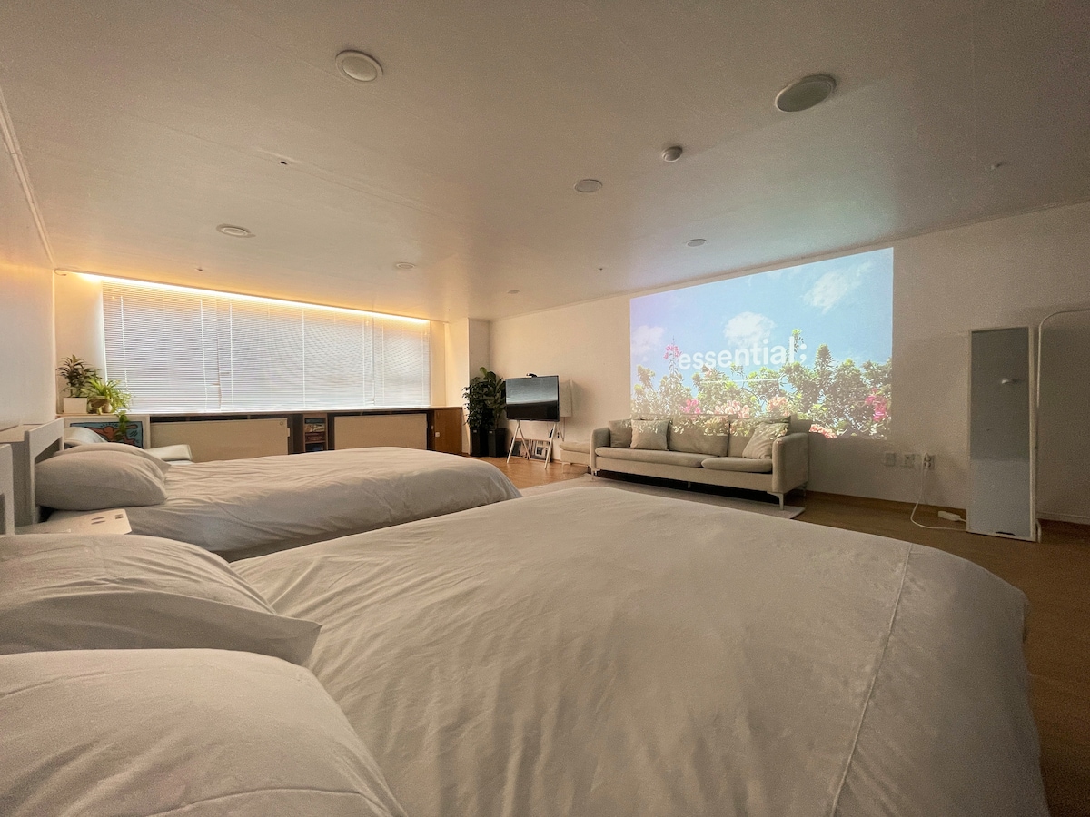 A spacious room is featuring two large beds with clean linens. A comfortable sofa is positioned in front of a wall that displays a large projection screen. Natural light enters through a broad window, creating a bright and inviting environment.