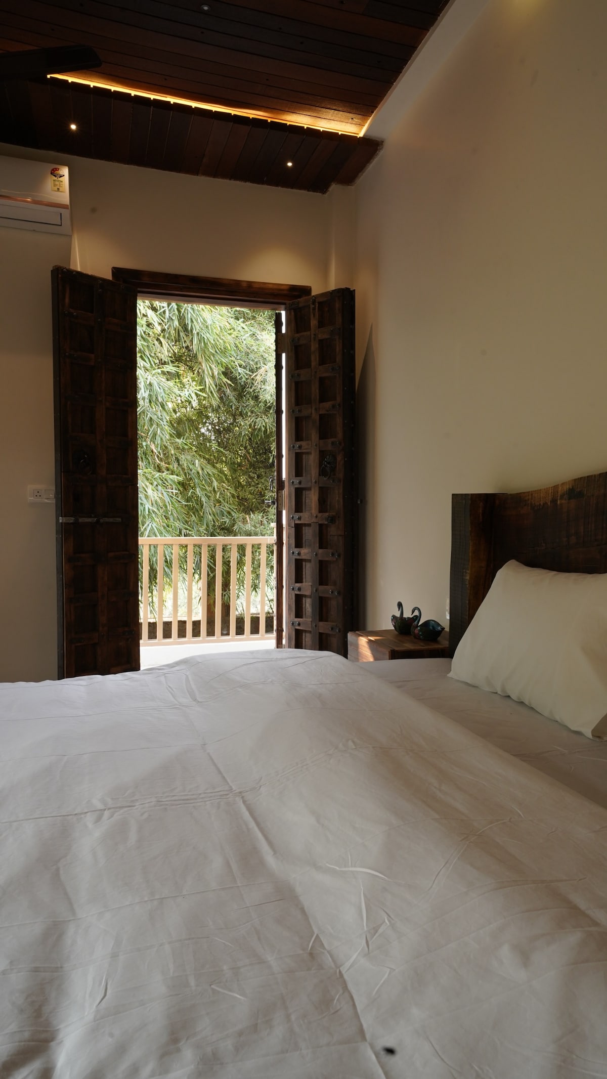 A cozy sleeping area features a spacious bed with a crisp white bedspread. Natural light filters through an open wooden door, revealing a view of lush greenery just outside. The room is softly illuminated, creating a calm and inviting setting.