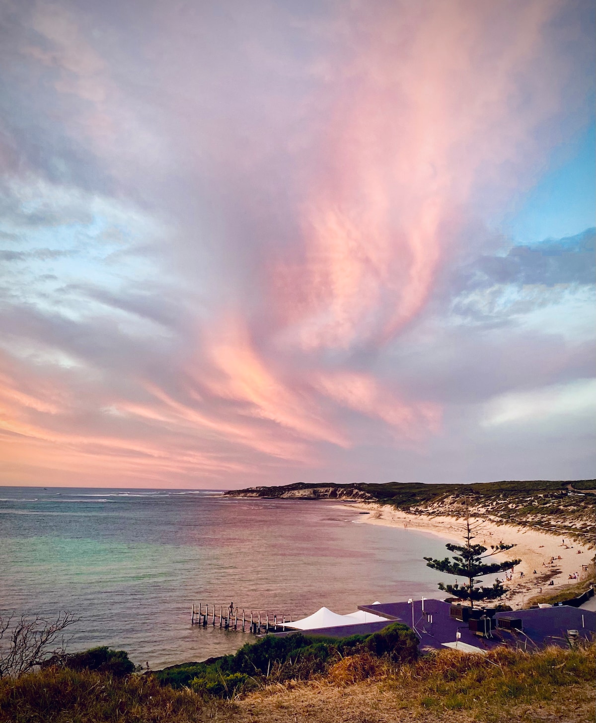 A serene coastal view is presented, showcasing a sandy beach that extends along the shoreline. The sky features soft pink and purple hues blending into blue, contrasting with the gentle waves of the ocean. Coastal vegetation and a distant structure are also visible.