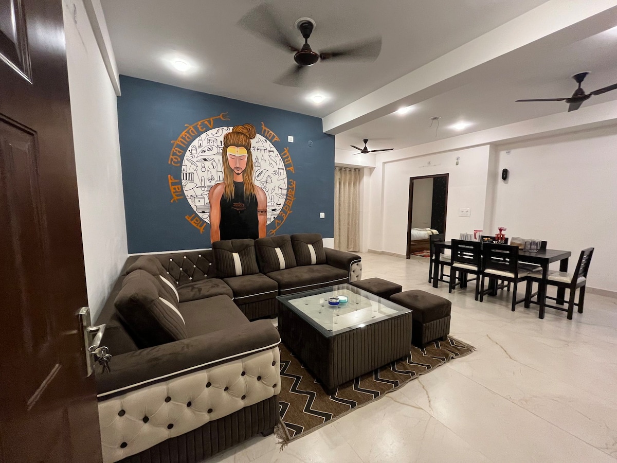 A modern living room showcases a spacious L-shaped sofa with plush cushions and a glass coffee table. A distinctive mural is featured on an accent wall, while a dining table and chairs are positioned nearby. Ceiling fans and ample natural light enhance the space.