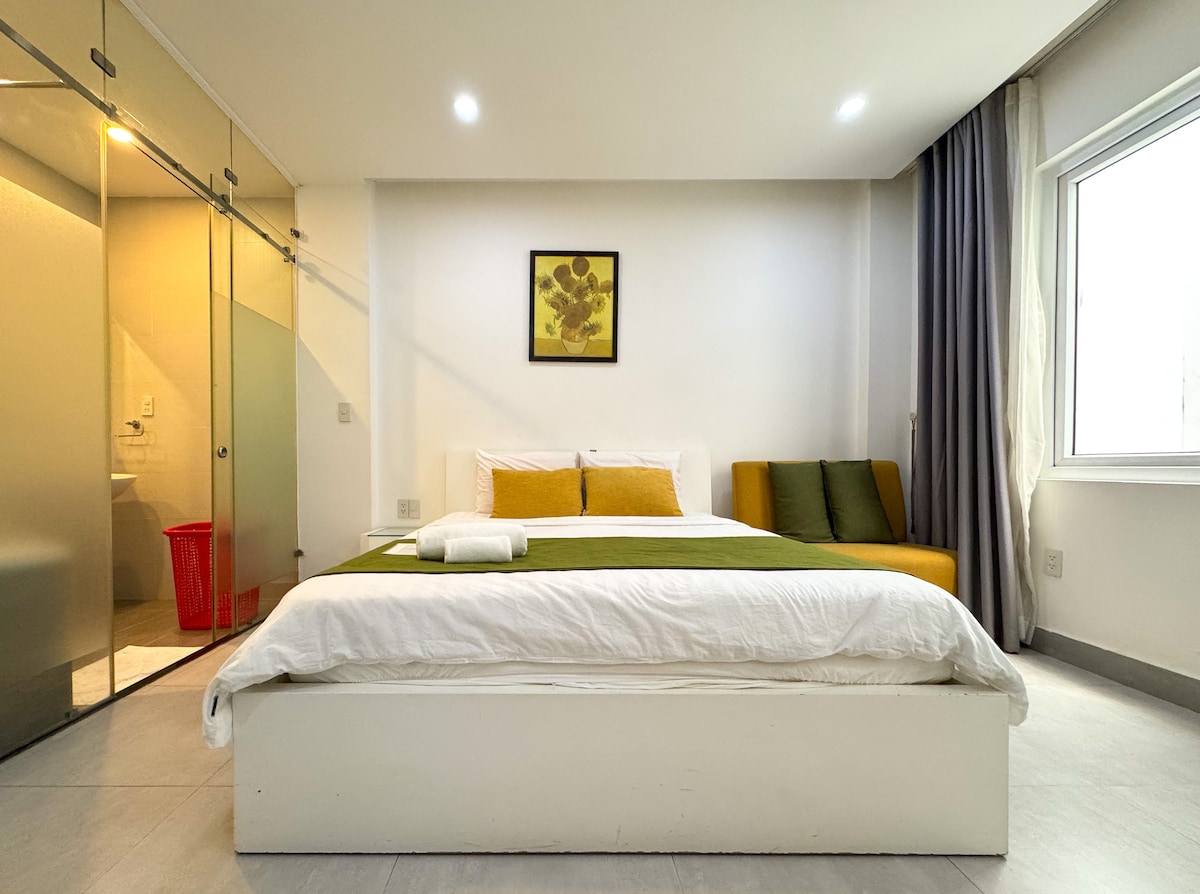 A modern bedroom is presented with a spacious bed featuring crisp linens and an accent pillow. A small seating area with green cushions is placed beside a window, while a glass partition reveals an ensuite bathroom. Soft lighting highlights the room's minimalistic design.