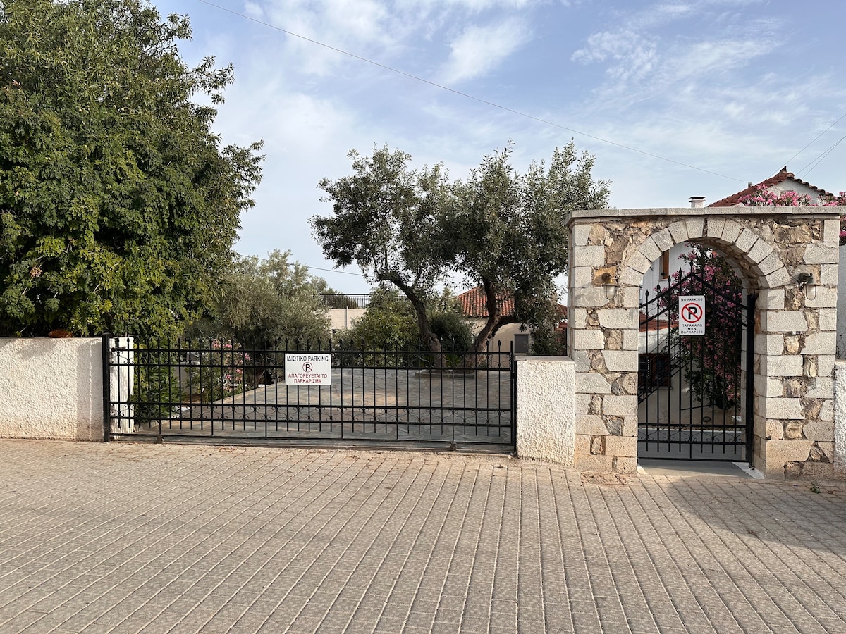 A gated entrance presents a stone archway, flanked by lush greenery and trees. The driveway leads into a spacious area, which is paved and includes a parking area marked with signage. A clear blue sky provides a bright backdrop.