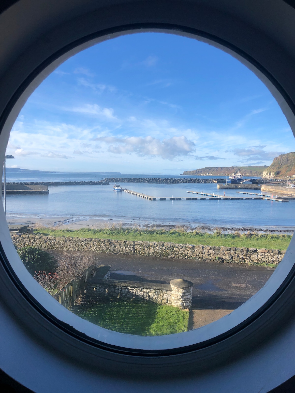 A round window offers a view of a calm harbor featuring boats moored against a backdrop of rolling hills. The sky is bright blue with scattered clouds, while the shoreline is lined with a stone wall and greenery in the foreground.