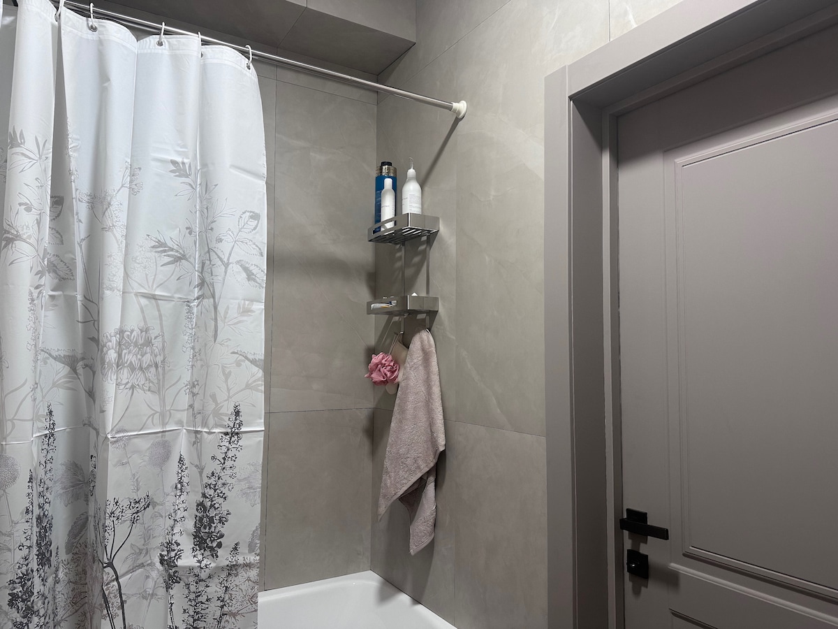 A modern bathroom is shown, featuring a soothing gray tile wall. A light-colored shower curtain with a floral design hangs, while a wall-mounted shelf accommodates toiletries. A plush towel is neatly hung nearby, with a door visible in the frame.