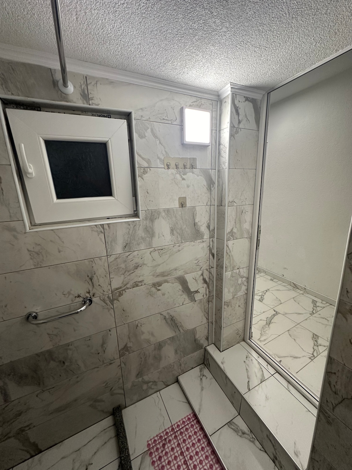 A modern bathroom features marble wall tiles and a large shower area. A mirror reflects the space, and natural light enters through a small window. A textured pink mat is placed on the floor, adding contrast to the light-colored tiles.
