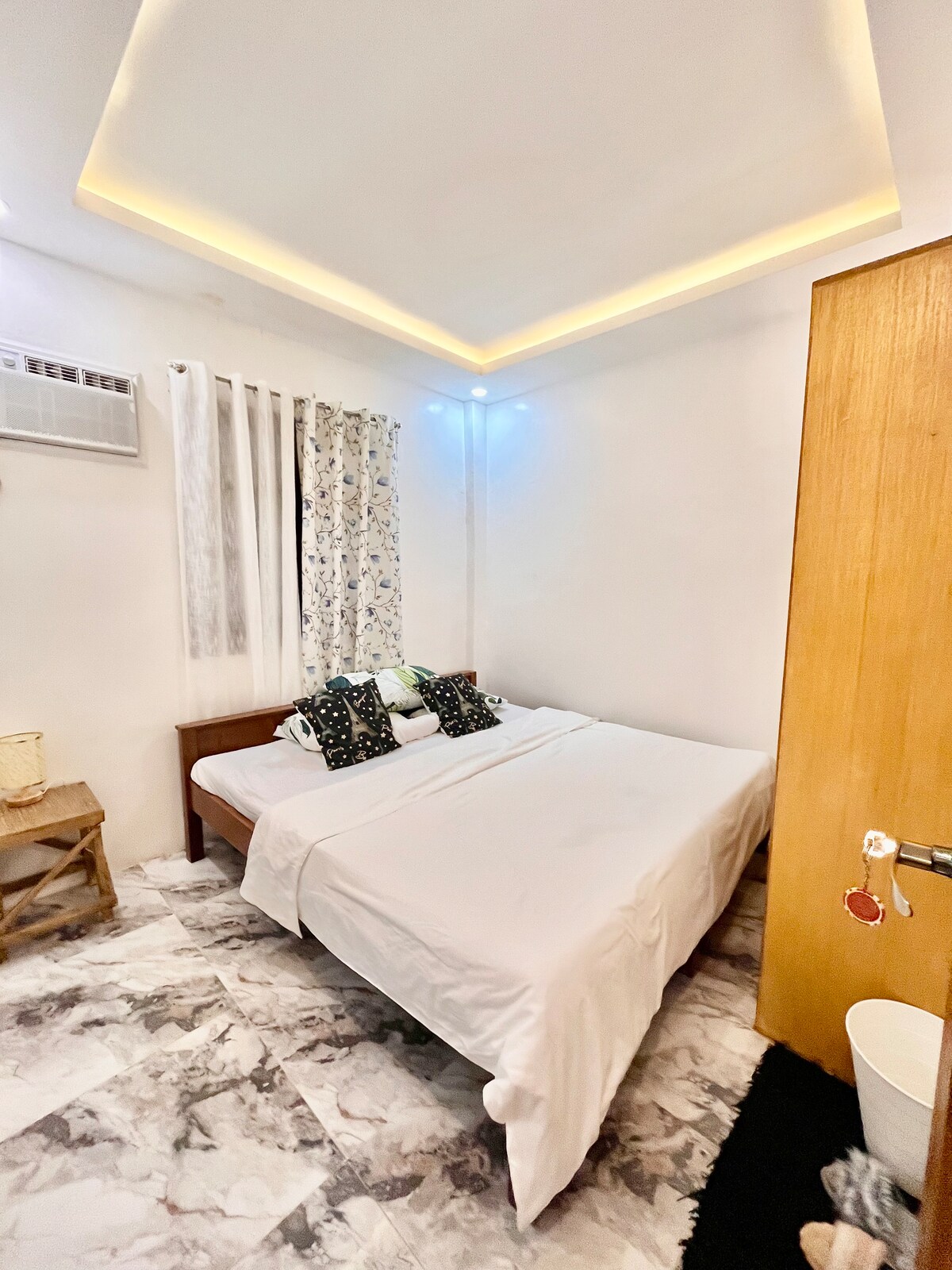 The sleeping area features a crisp white bedspread on a spacious bed, complemented by decorative pillows. Natural light filters through floral curtains, while a wooden door and a small side table add functional charm to the room's minimalistic design.