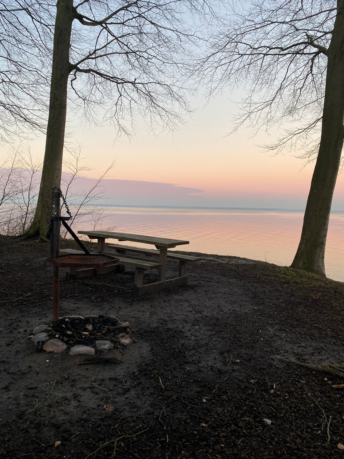 A serene lakeside view is captured at dusk, featuring a wooden picnic table positioned near the water. A fire pit with stones surrounds a circle of ash, while trees stand tall along the shoreline, creating a peaceful atmosphere by the calm water.