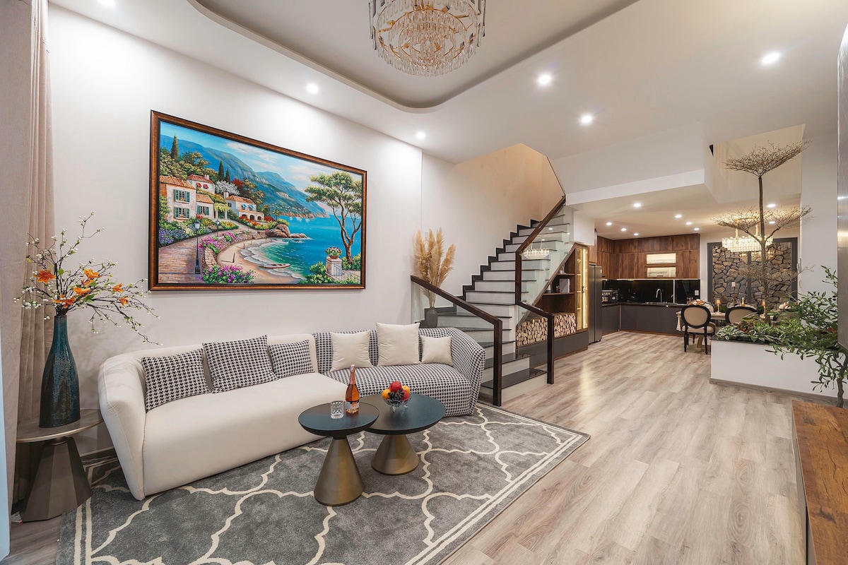 A modern living area features a light-colored sofa paired with two circular tables, complemented by decorative elements such as a large painting and floral arrangements. A staircase leads to the upper level, while the kitchen area and dining space are visible in the background.