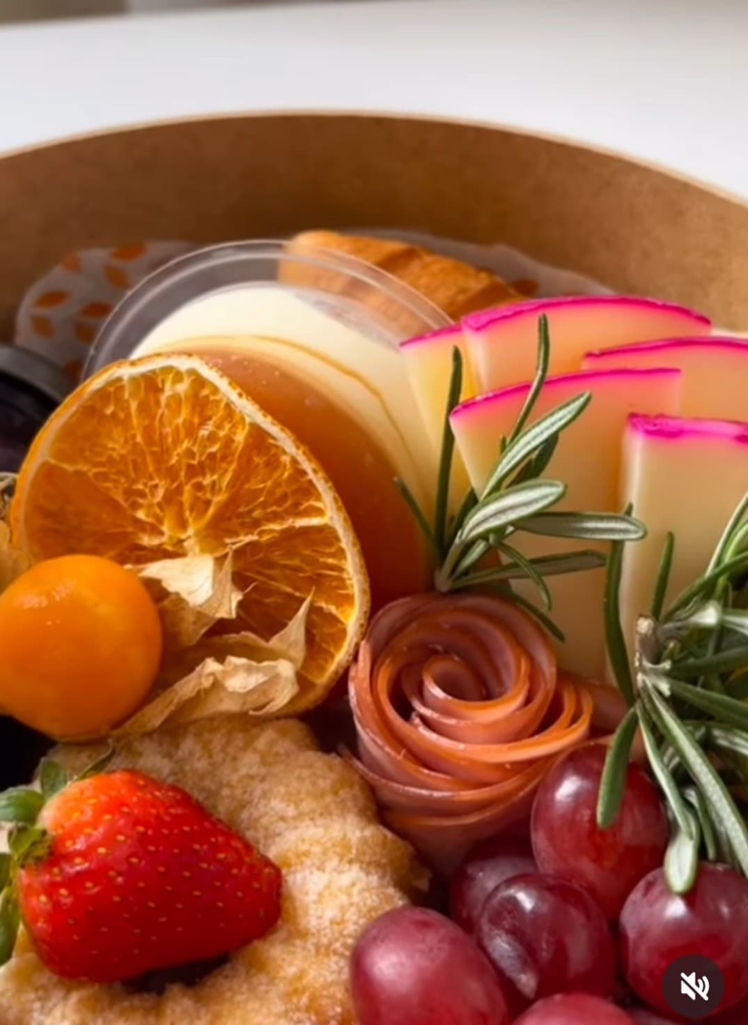 An assortment of fresh and dried fruits is displayed, featuring vibrant strawberries, grapes, and orange slices. A creamy dessert, rolled meats, and a croissant are included, all arranged in an inviting manner. Rosemary sprigs add a touch of greenery to the presentation.