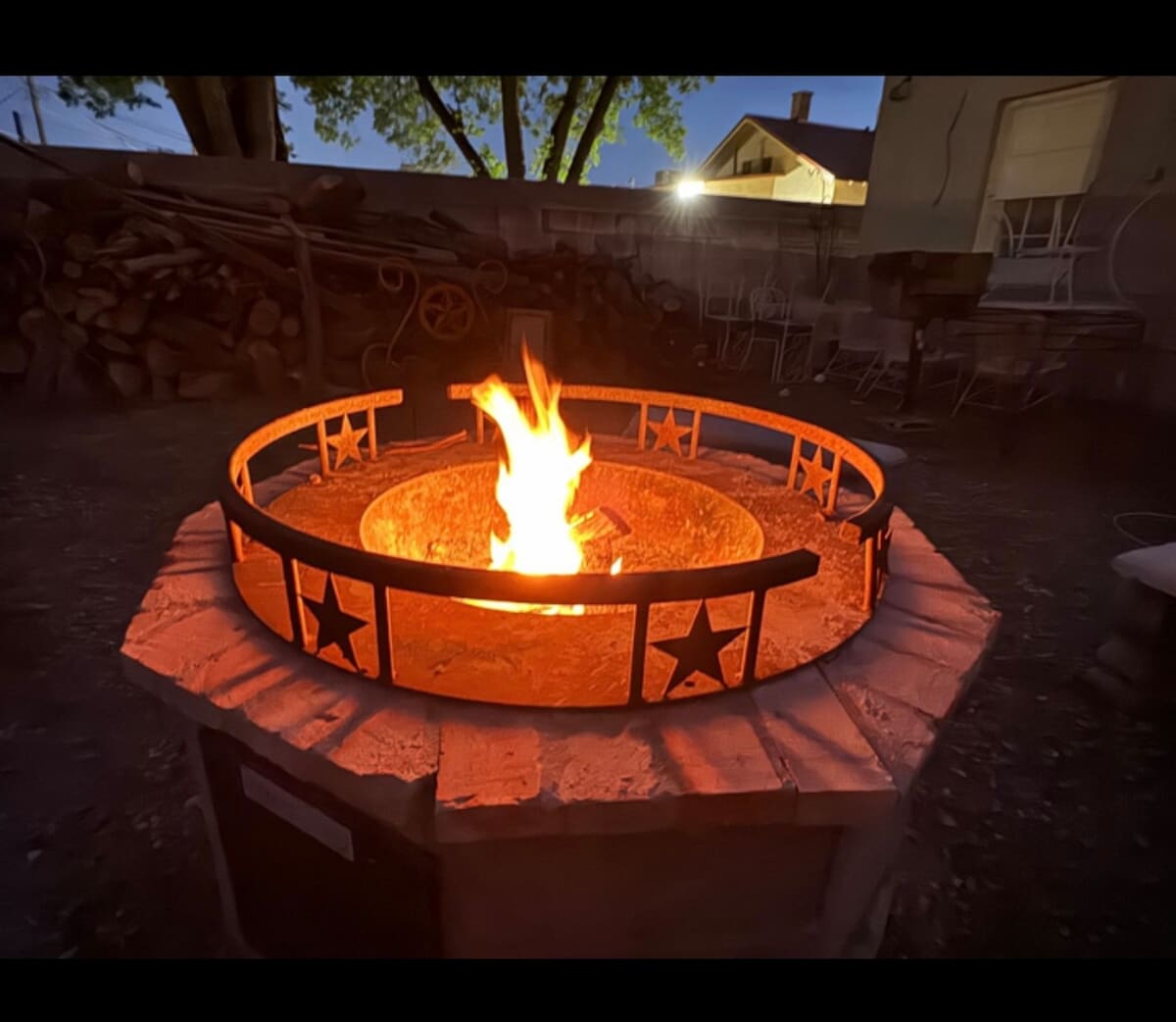 A circular fire pit is positioned at night, featuring a stone enclosure and a metal railing with star motifs. The flames create a warm glow, illuminating the surrounding area. In the background, dimly lit structures and firewood can be seen, enhancing the outdoor atmosphere.