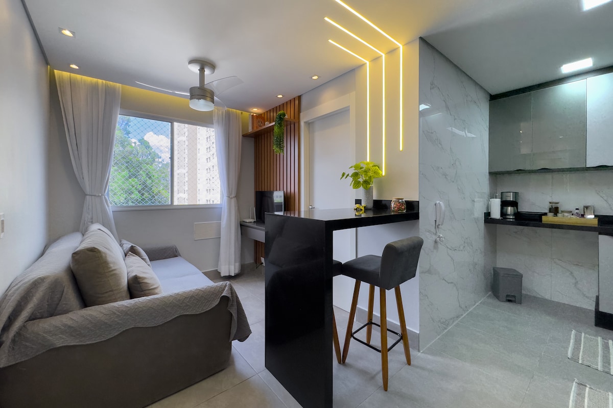 An inviting living space features a cozy sofa positioned near a large window that offers natural light. A compact kitchen area with modern finishes can be seen, complete with a countertop and barstool. Soft lighting accents give a warm ambiance to the room.