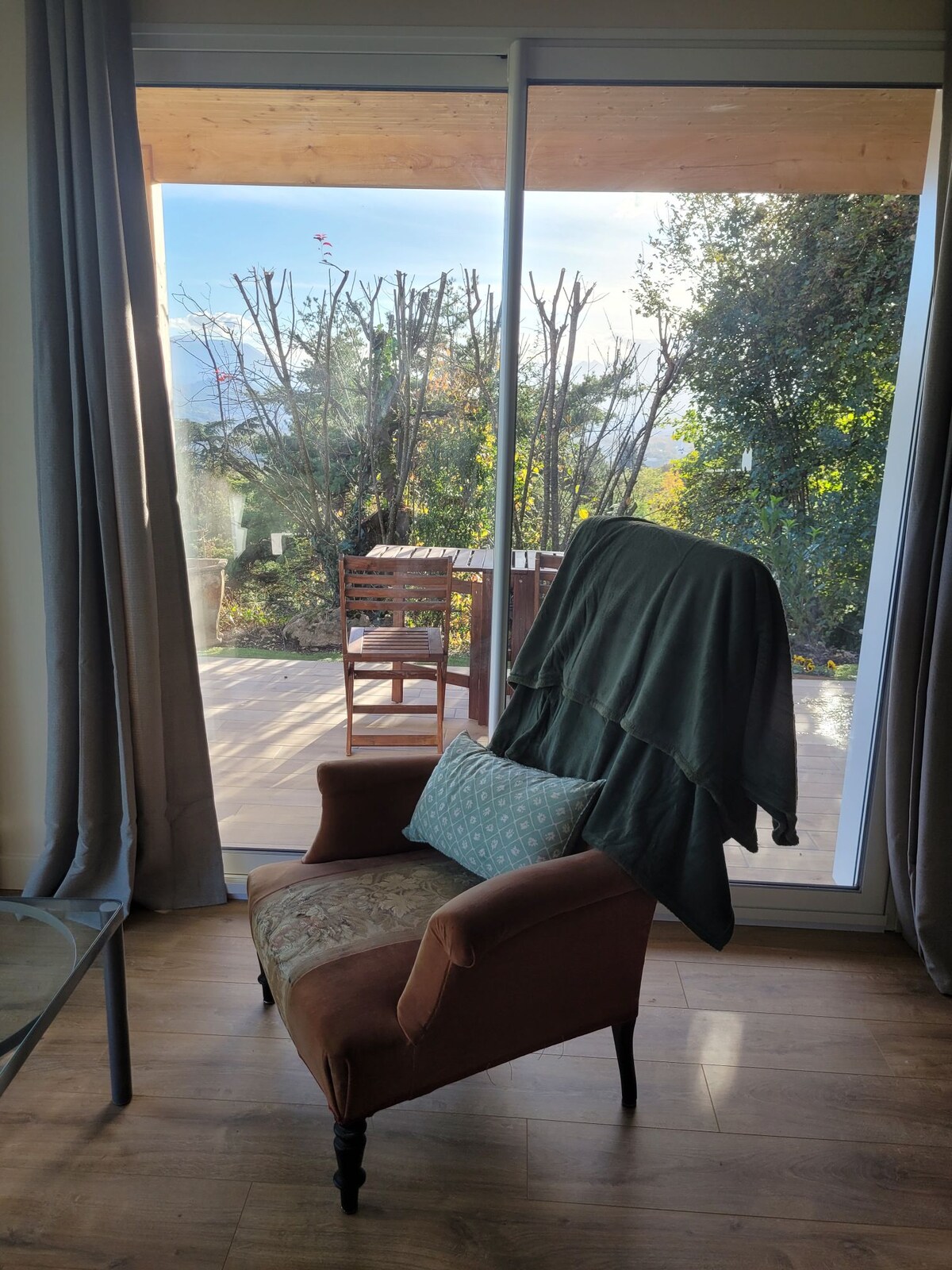 A cozy armchair with a green throw draped over it is positioned near large glass doors, leading to a wooden patio. Sunlight filters through, illuminating the space, while greenery and seating are visible outside.