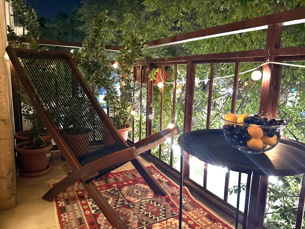 A cozy balcony is furnished with a wooden lounge chair and a round table. A bowl of fruits, including black grapes and oranges, is placed on the table. Soft lighting and greenery from plants create a welcoming atmosphere. A colorful rug enhances the outdoor space.
