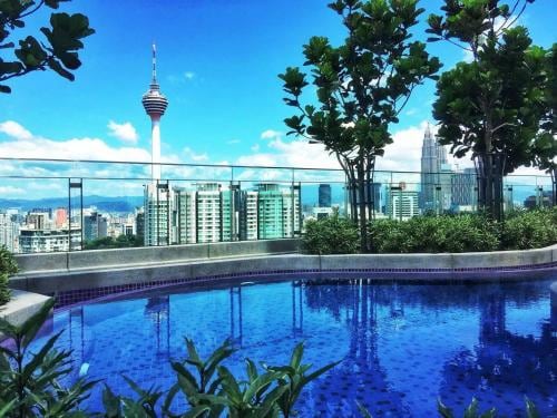 A rooftop infinity pool is framed by lush greenery, providing a view of the Kuala Lumpur skyline including the iconic KL Tower. The serene blue water reflects the clear sky, creating a tranquil atmosphere ideal for relaxation.