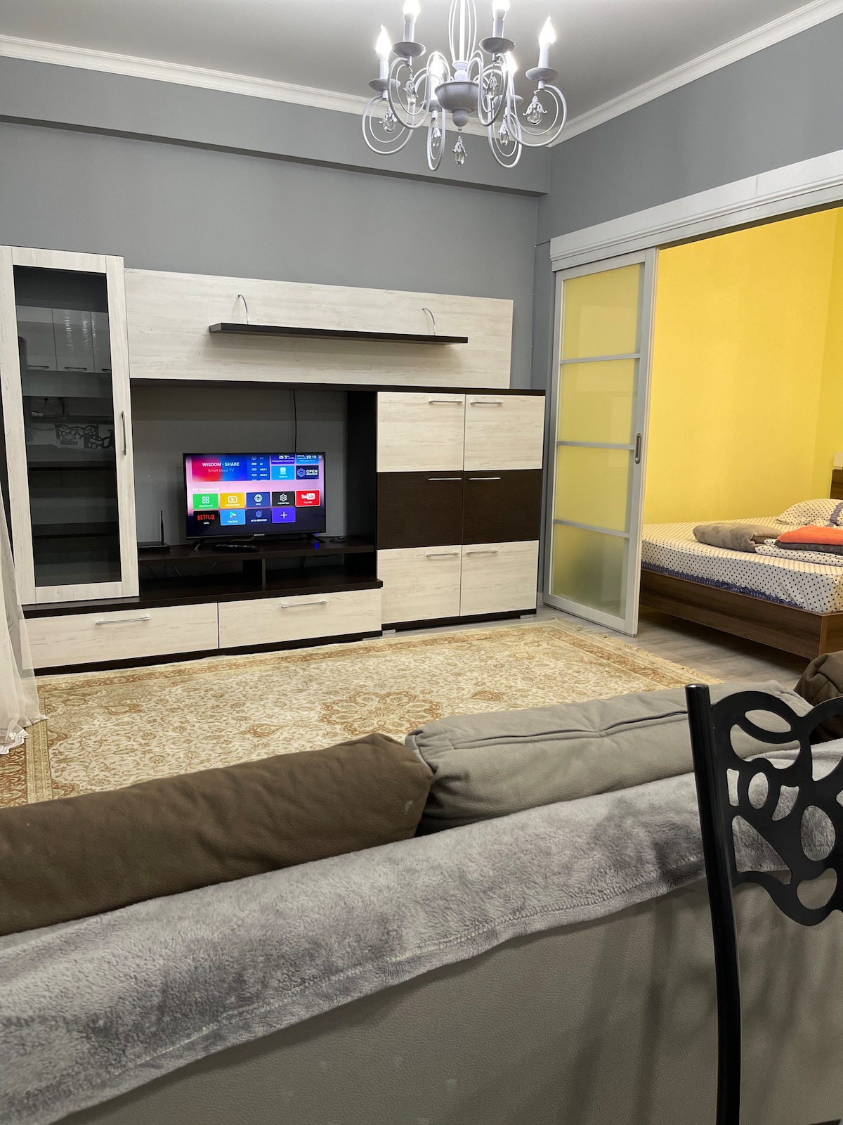 A modern living area features a sleek entertainment unit with a flat-screen TV displaying various apps. Light-colored furniture is arranged on a decorative area rug. A cozy sleeping area is visible through glass doors, with soft bedding and a vibrant yellow wall providing a contrasting backdrop.