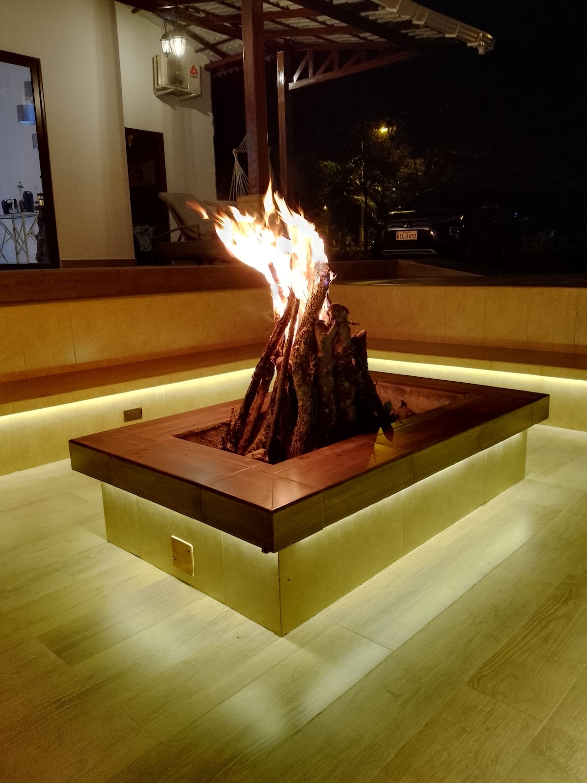 A wooden fire pit is centered on a warm wooden platform, surrounded by soft, illuminated edges. Flames dance atop a stack of logs, providing a cozy ambiance. The area is framed by smooth wooden seating that enhances the inviting environment.