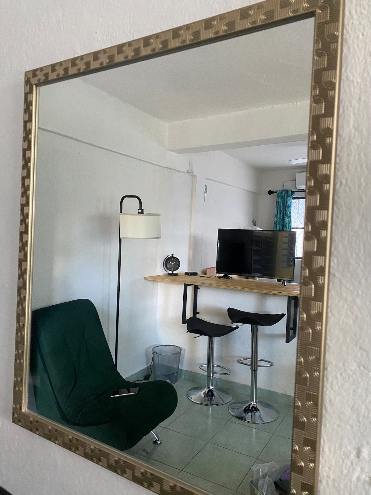 A wall-mounted mirror reflects a cozy seating area, highlighting a green chair and a small wooden table with two bar stools. A lamp stands beside the chair, and a television is positioned on a nearby surface, contributing to the functional layout of the space.