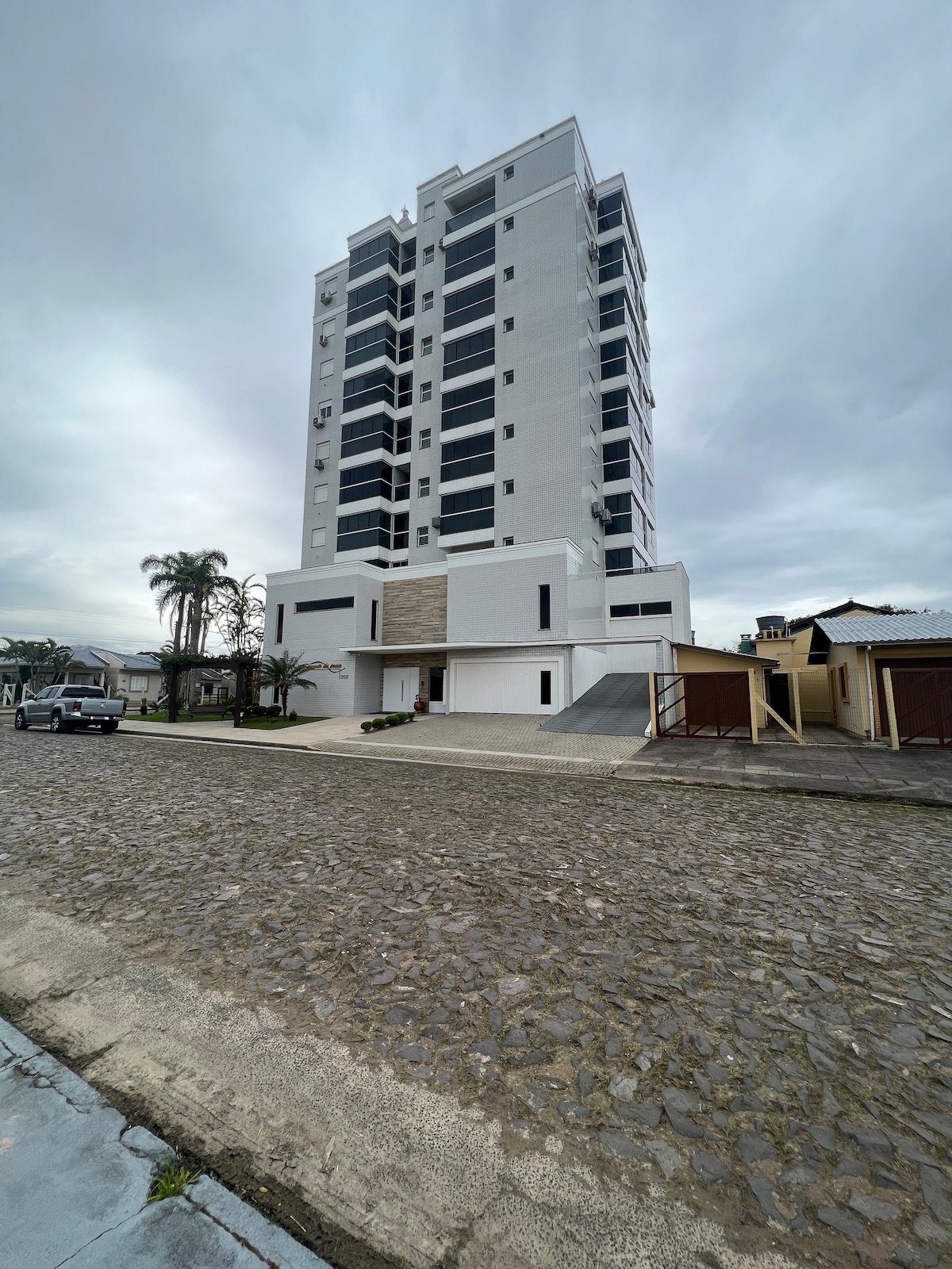 A modern multi-story building stands prominently along a cobblestone street, showcasing a sleek white facade. Large windows create an open appearance, while palm trees line the entrance, adding greenery to the urban setting. The structure includes a covered driveway leading to the entrance.
