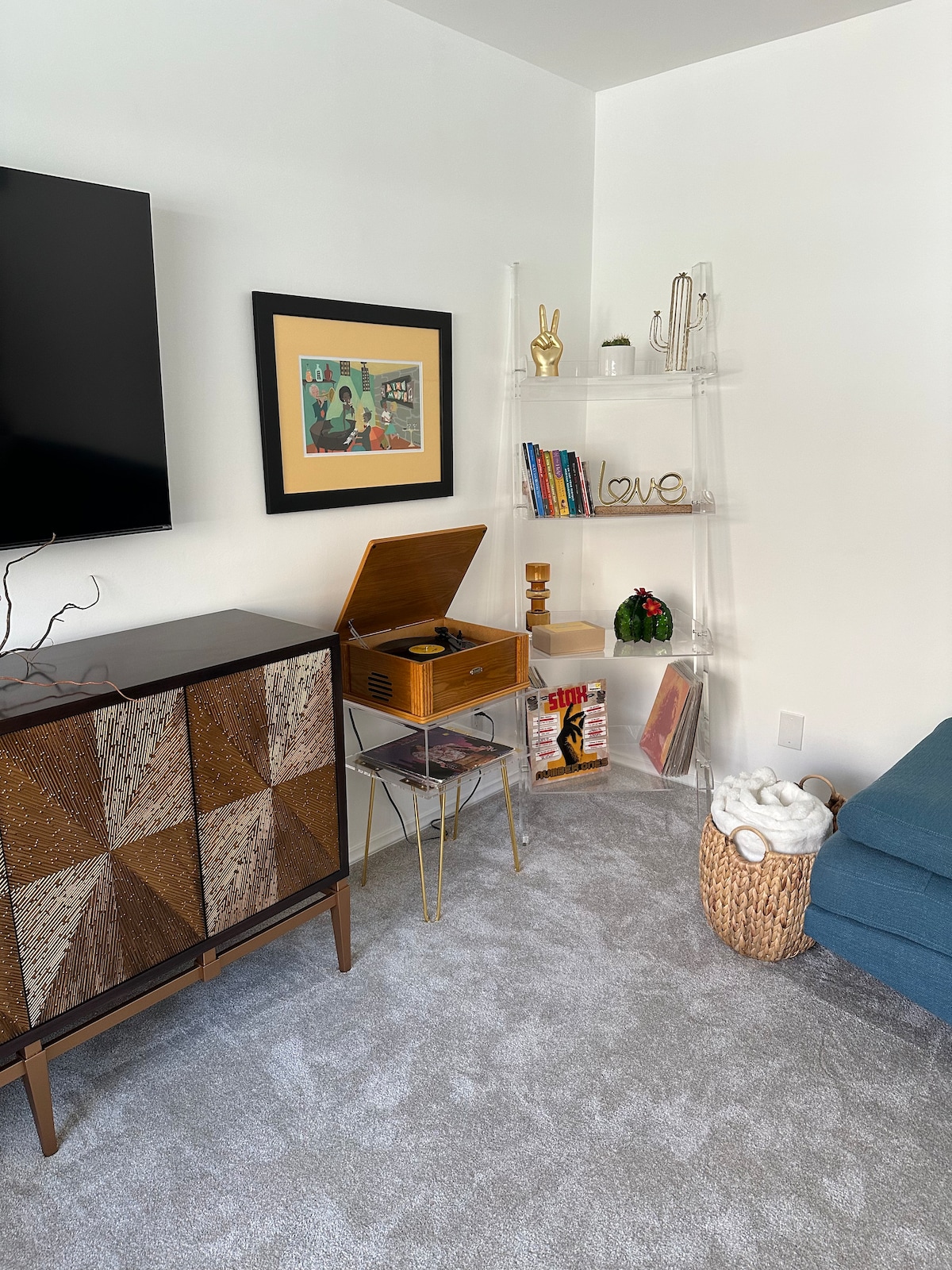 A cozy living room corner features a retro record player atop a glass table, surrounded by decorative accents. A stylish cabinet displays geometric patterns, while a nearby shelf holds books and plants, contributing to a relaxed atmosphere with a plush blue sofa nearby.