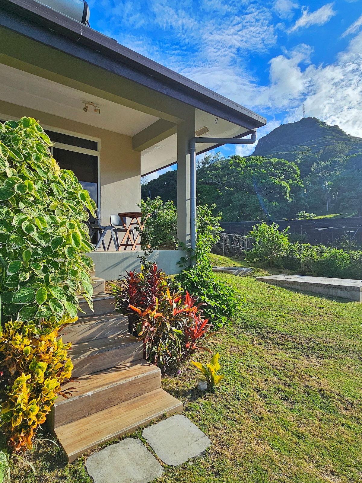 The bungalow entrance is surrounded by lush greenery and colorful plants. Steps lead up to a welcoming porch that offers views of Mount Tapioi in the distance. The bright sky complements the vibrant colors of the garden, creating a serene outdoor space.