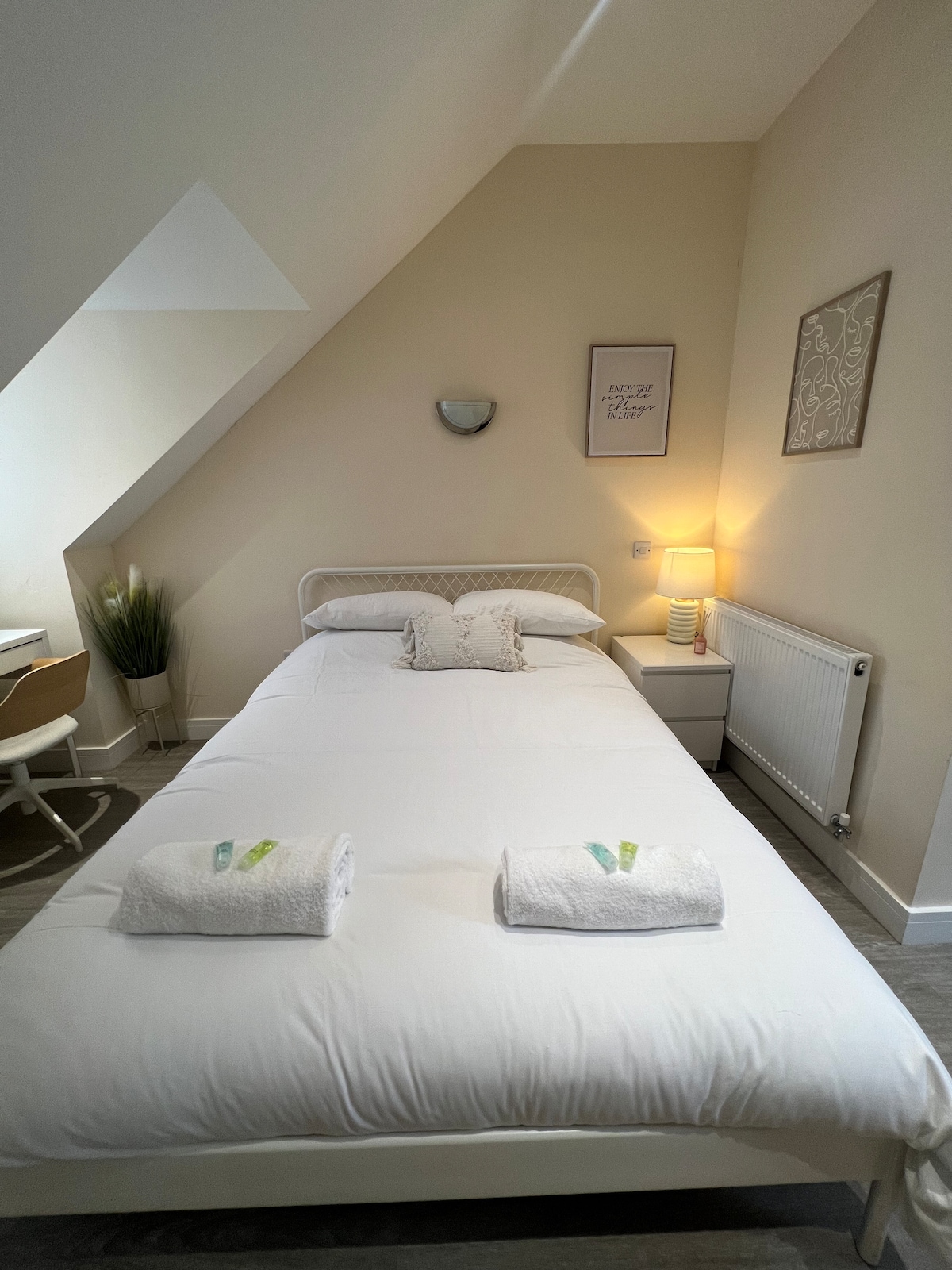 A well-lit sleeping area features a spacious bed with neatly arranged towels at its foot. A bedside table with a lamp sits beside the bed, while a small desk and chair are positioned near a window. Decorative plants and wall art enhance the space.