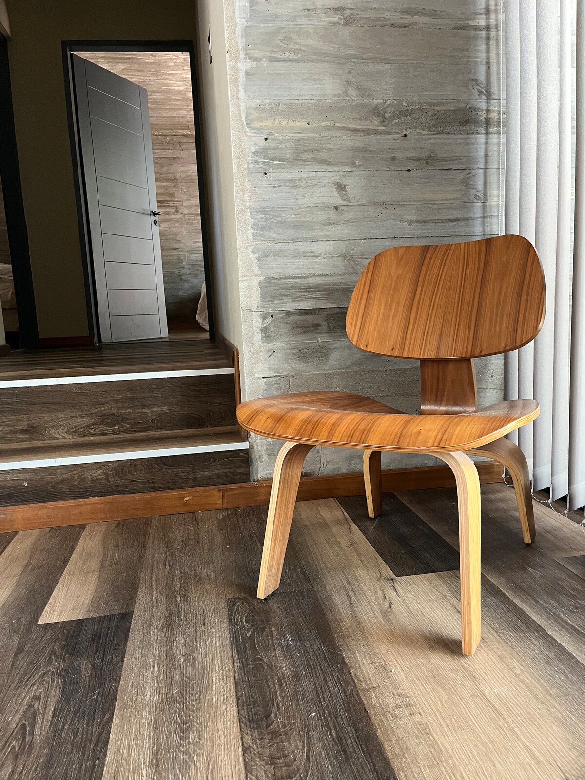 A wooden chair with a curved backrest is positioned next to a staircase. Light filters through vertical blinds, highlighting the chair’s natural finish. The floor features a mix of wood tones, creating a warm ambiance, while a partially visible door adds to the interior's depth.