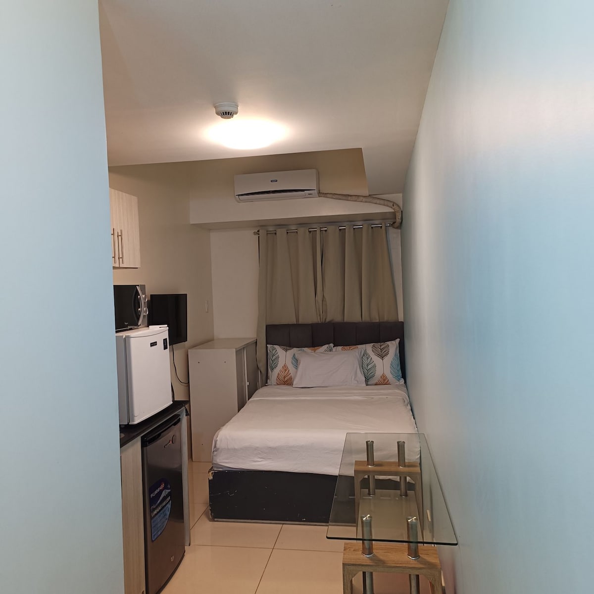 A compact room features a spacious bed with decorative pillows. Light walls create a bright environment. A small table stands beside the bed, while a kitchenette with a microwave and refrigerator is nearby. Curtains frame a window, allowing natural light into the space.