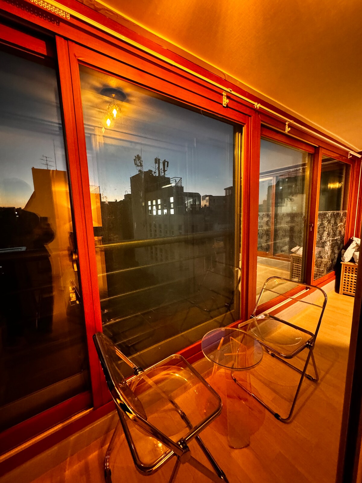 A small balcony area features two transparent chairs positioned beside a glass table. Floor-to-ceiling windows provide a view of the cityscape, with warm sunset hues reflecting off the glass. Soft lighting enhances the ambiance, creating a cozy space.