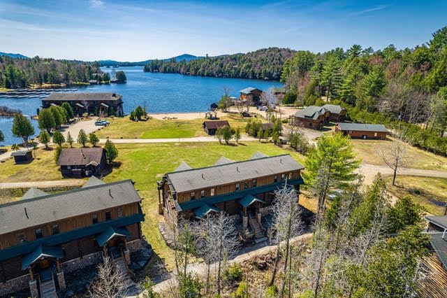 An aerial view captures serene waters and lush greenery surrounding the cabin area. The spacious sandy beach along Ampersand Bay is visible, bordered by well-maintained grounds and scattered structures. Mountains rise in the distance, enhancing the natural beauty of the location.
