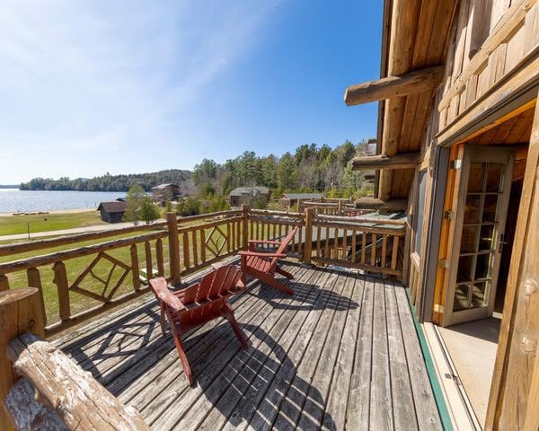 A spacious deck showcases wooden lounge chairs resting on a natural wood surface, with a view of a tranquil lake and distant trees under a clear blue sky. The cabin's French doors provide an entryway to the indoor space.