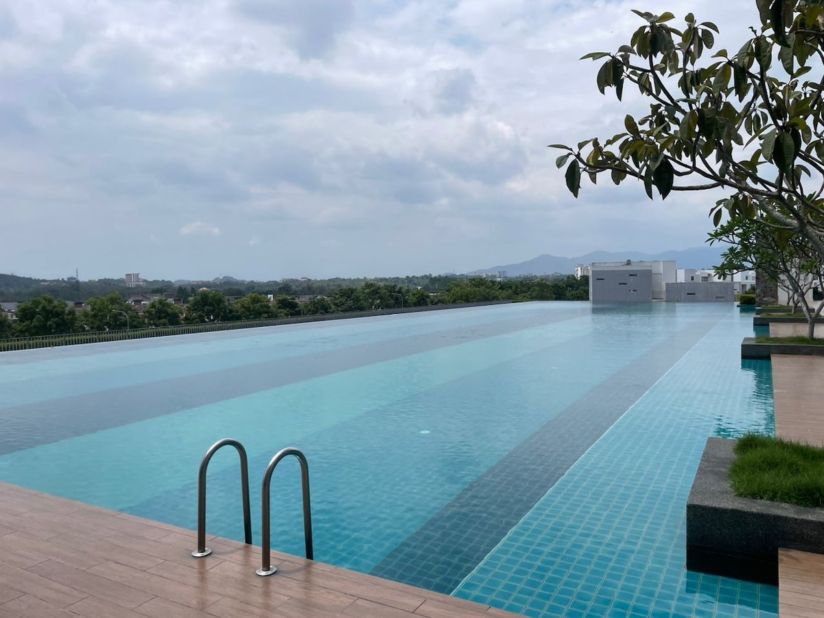 An infinity pool stretches across the image, featuring a smooth water surface reflecting the sky. Surrounding greenery adds a natural touch, while gentle hills are visible in the distance. The area is designed for relaxation, with pool access indicated by steps leading into the water.