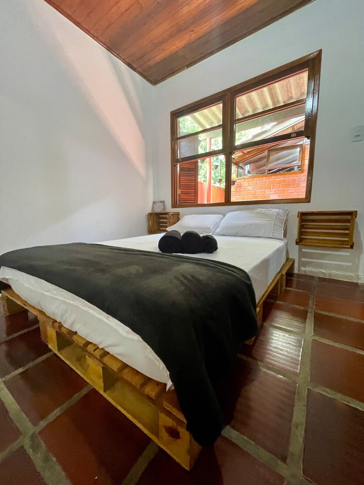 A cozy sleeping area features a spacious bed with a black throw blanket and two pillows. Natural light enters through a large window, which overlooks the exterior greenery. Side tables are positioned alongside the bed, complemented by a rustic wood floor.