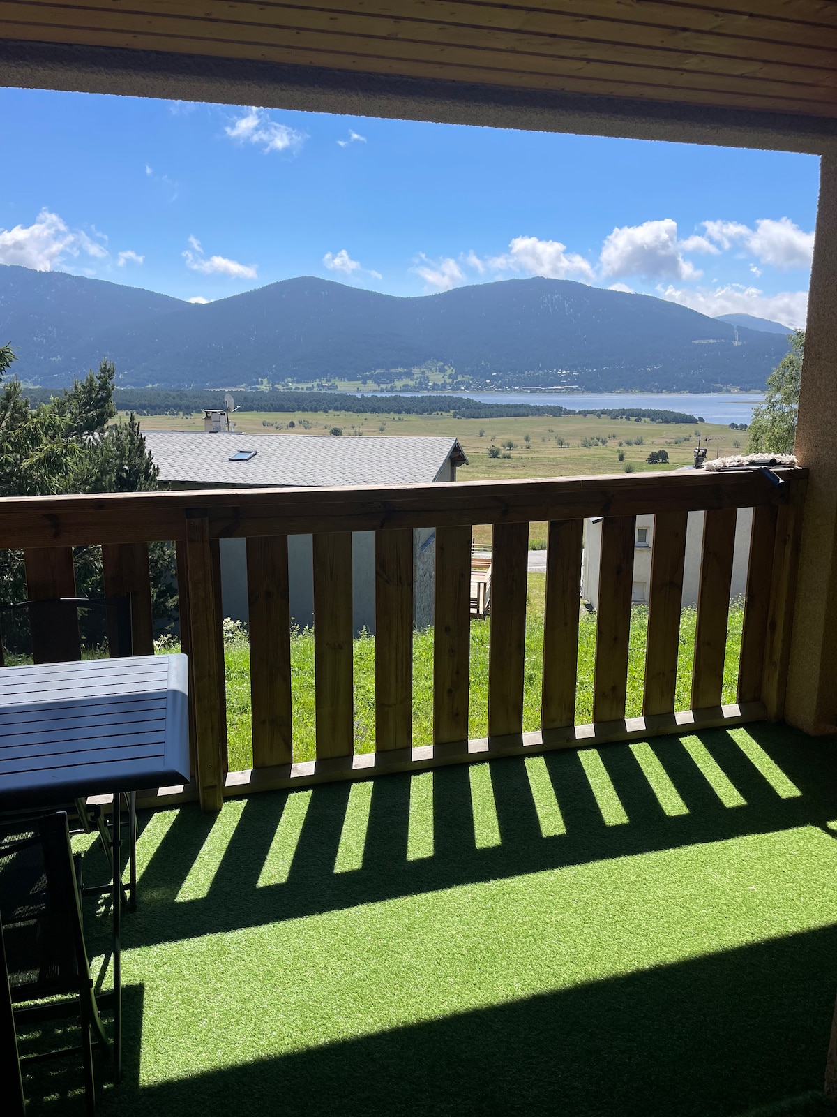 A balcony offers a sweeping view of the surrounding mountains and lake. Beneath a clear blue sky, sunlight casts shadows on the green artificial turf. Simple outdoor furniture is visible, providing a space for relaxation while enjoying the scenic landscape.