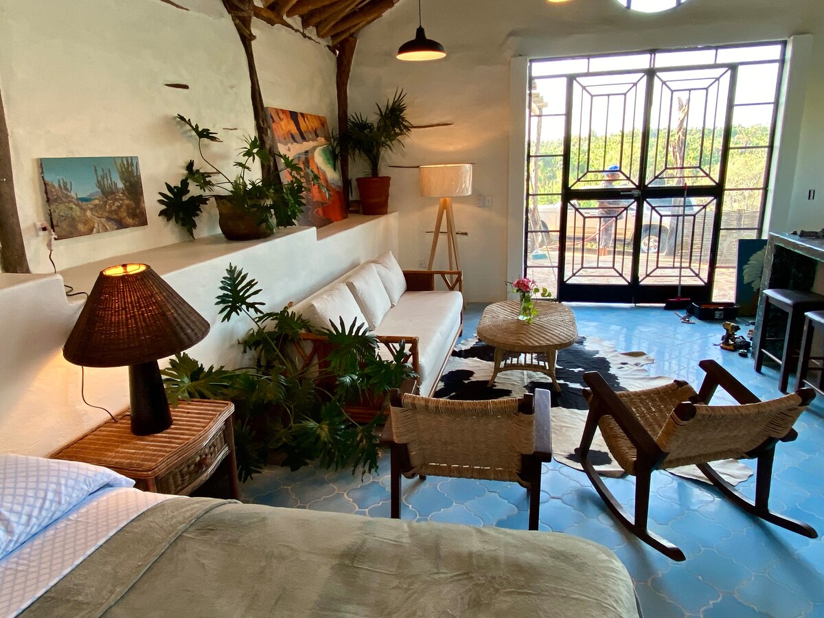 A cozy living space is highlighted by a light-colored sofa, a wooden rocking chair, and a decorative coffee table. Lush plants enhance the ambiance, while large windows provide natural light and a view of the outdoors. A textured lamp adds warmth to the area.