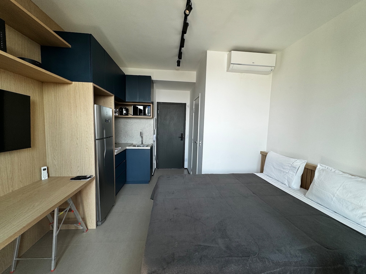 A modern studio apartment features a spacious bed with soft linens, complemented by a sleek kitchenette and dining area. Light wood cabinetry contrasts with dark blue furnishings. An air conditioning unit is mounted above, and a large window offers ample natural light in the minimalist space.