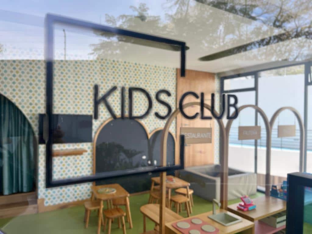 A vibrant kids' club area is visible, featuring playful decor with a cheerful backdrop. Small wooden tables and chairs are arranged for group activities. The space is designed with archways and colorful elements, providing a welcoming environment for children.