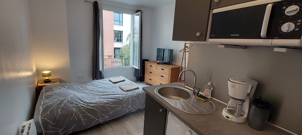 The studio features a spacious sleeping area with a bed covered in a textured gray blanket. A kitchenette with modern appliances is present, while a small table and TV are visible. Natural light enters through the large window, complemented by light curtains.