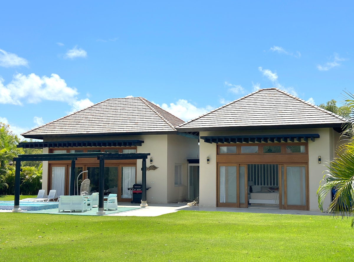 A spacious outdoor area features two well-appointed houses, each with large glass doors that open to a verdant lawn. The roofs have a traditional design, and a grill is positioned discreetly on the patio, complementing a serene environment.