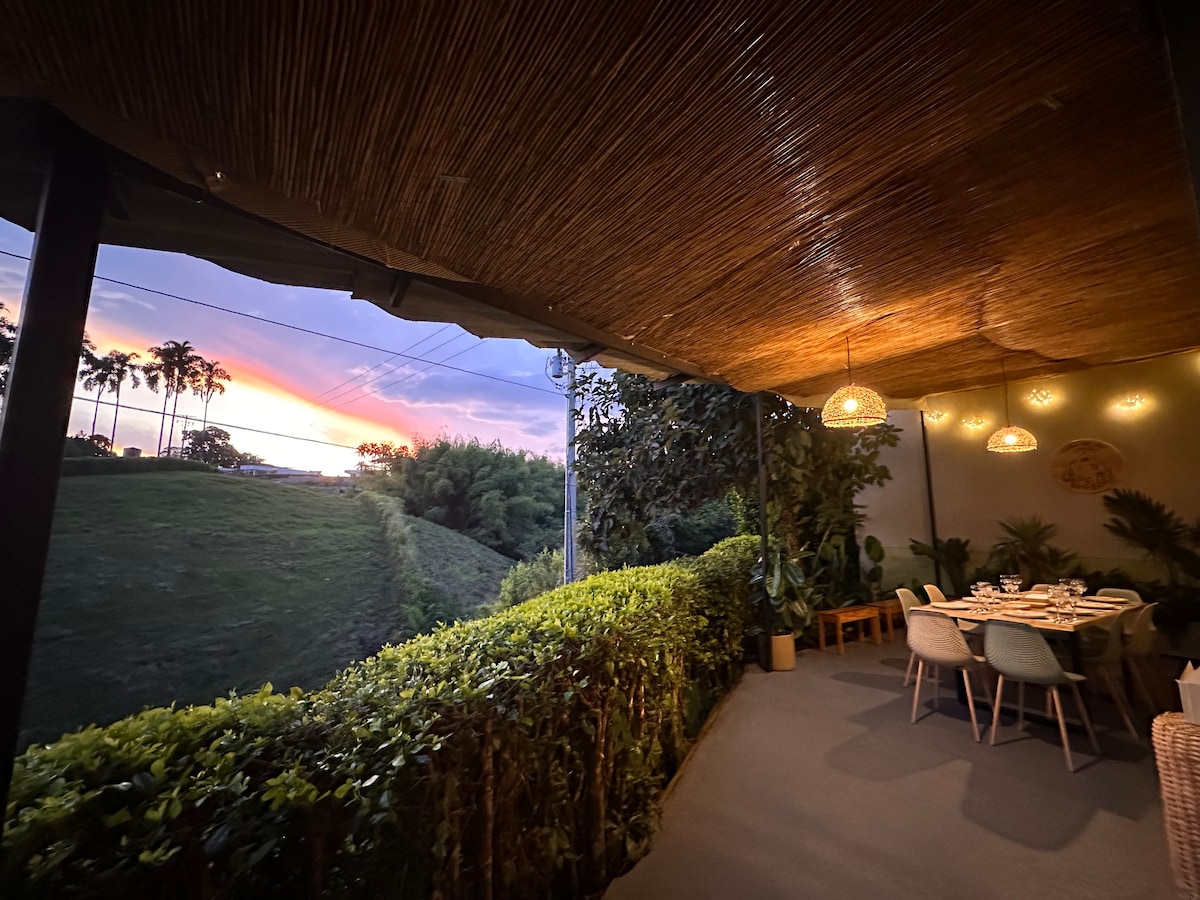 An outdoor dining area is presented with a view of a vibrant sunset, showcasing shades of pink and purple in the sky. Natural decor with greenery enhances the space, while pendant lighting adds a soft glow, creating a welcoming atmosphere for meals.