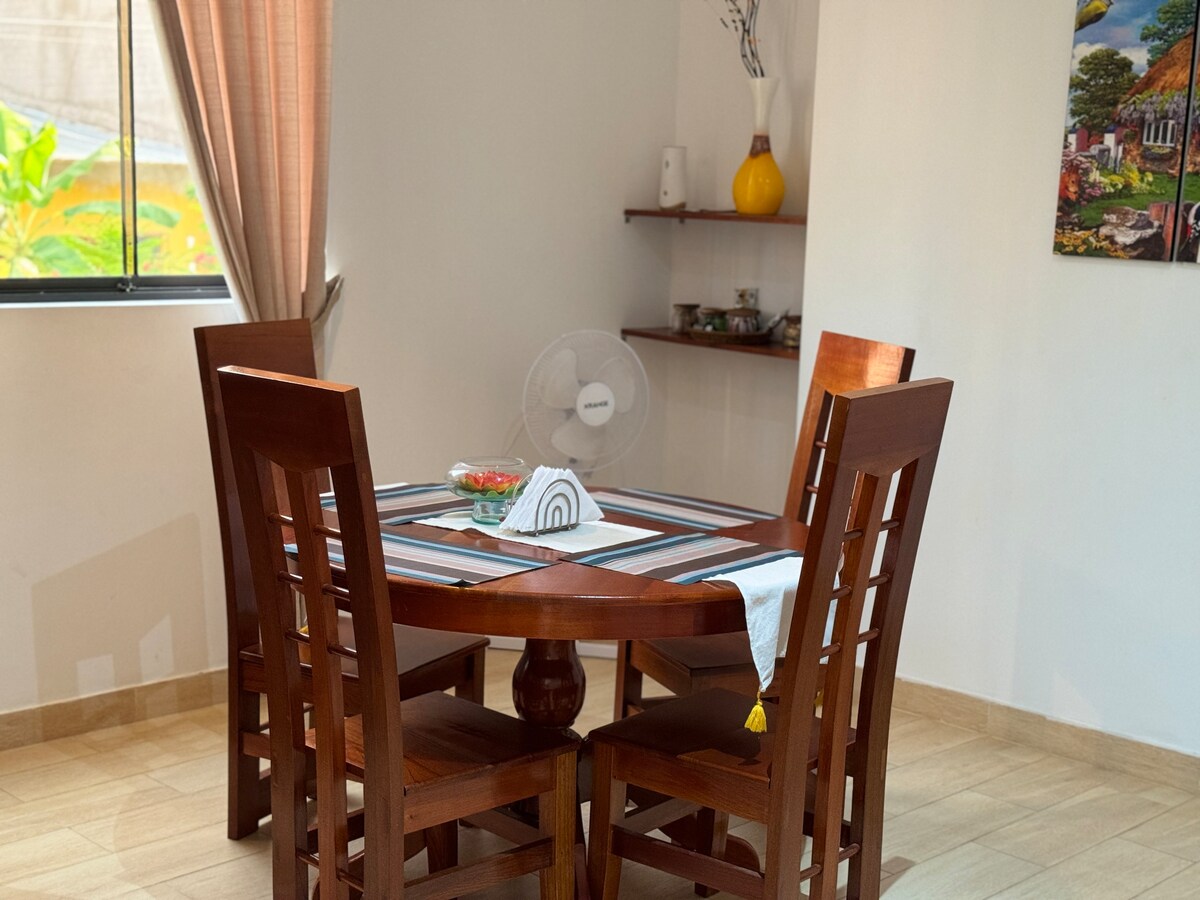 A wooden dining table is surrounded by four matching chairs, positioned near a window that allows natural light to fill the space. A ceiling fan is present to enhance airflow, while a colorful painting adorns the wall, adding visual interest.
