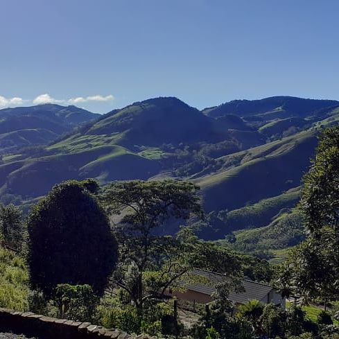 Lush green hills and rolling mountains create a serene landscape. The clear sky enhances the natural beauty of the surroundings, with trees dotting the foreground and a distant view of a nearby structure nestled among the hills.