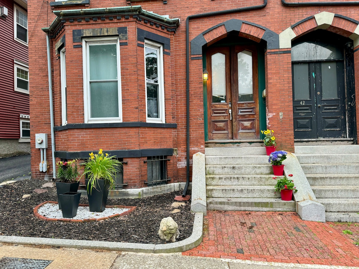 The entrance features two wooden doors with decorative arches, flanked by potted flowers in vibrant colors. Stone steps lead up to the doors, with a landscaped area at the base, showcasing mulch and a mix of greenery.