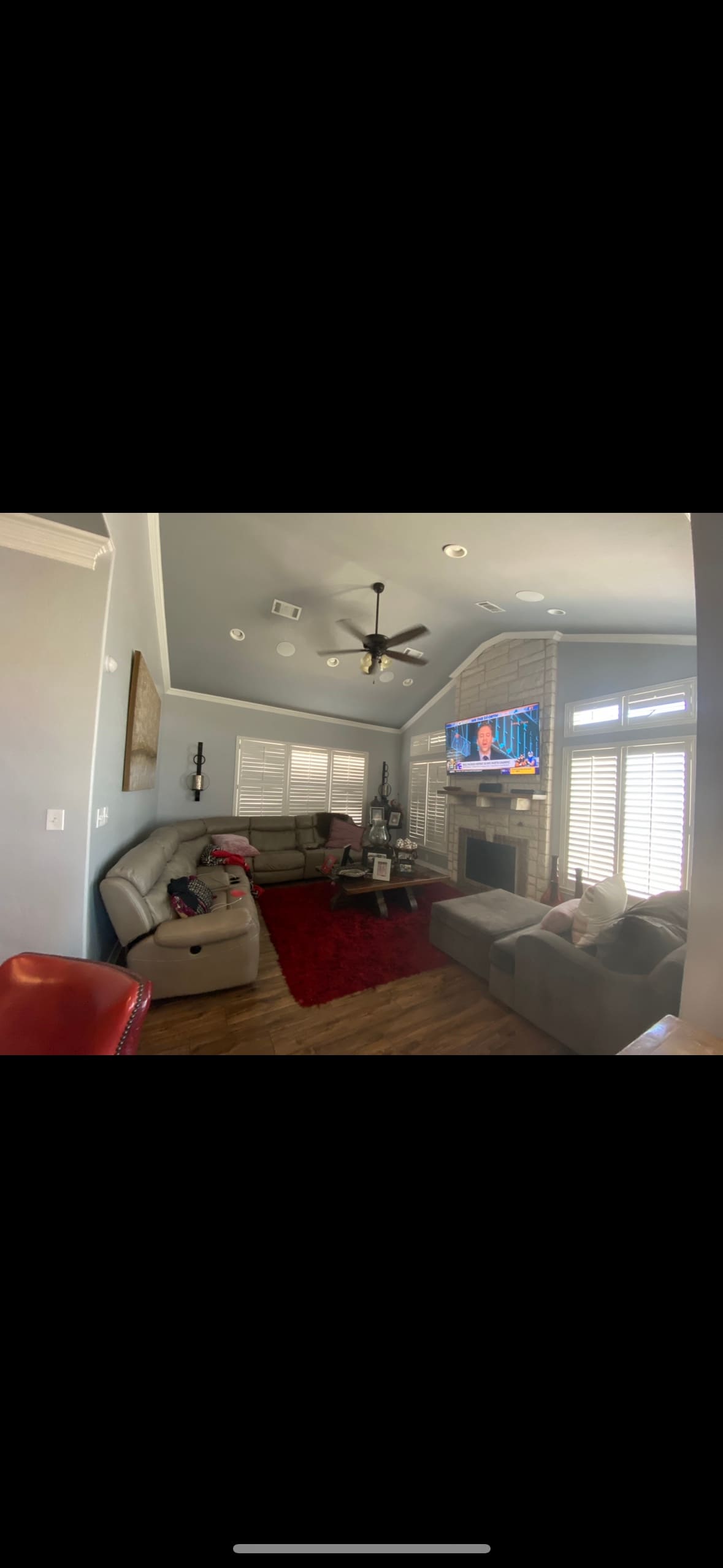 A cozy living room features a comfortable sectional sofa and a bright accent chair on warm wooden flooring. A red area rug adds a pop of color, while large windows are dressed with shutters, allowing natural light to fill the space. A television is mounted on the wall.