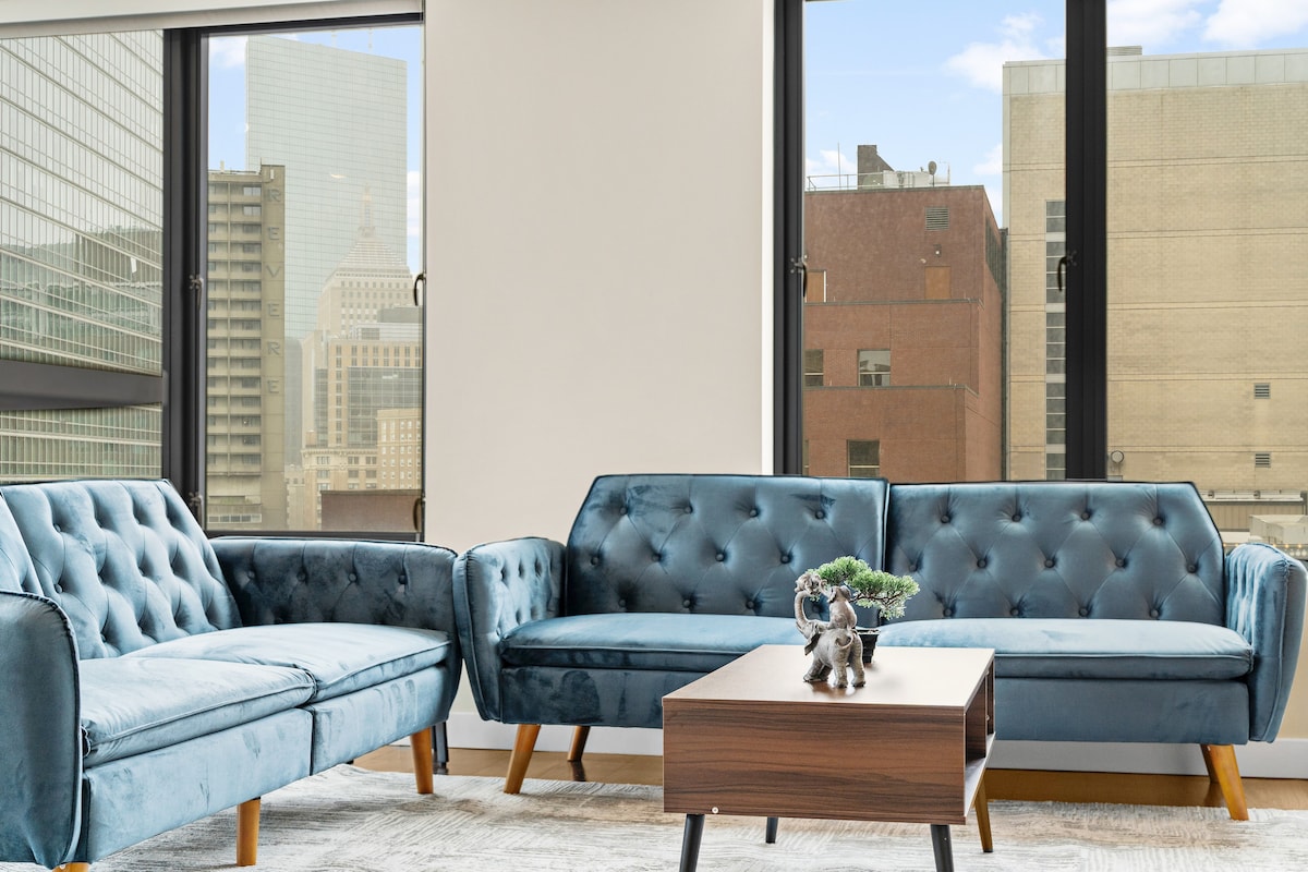 A modern living area features a pair of plush blue sofas positioned around a low wooden coffee table. Large windows allow natural light to fill the space, offering views of the urban skyline. A small decorative plant rests on the table.