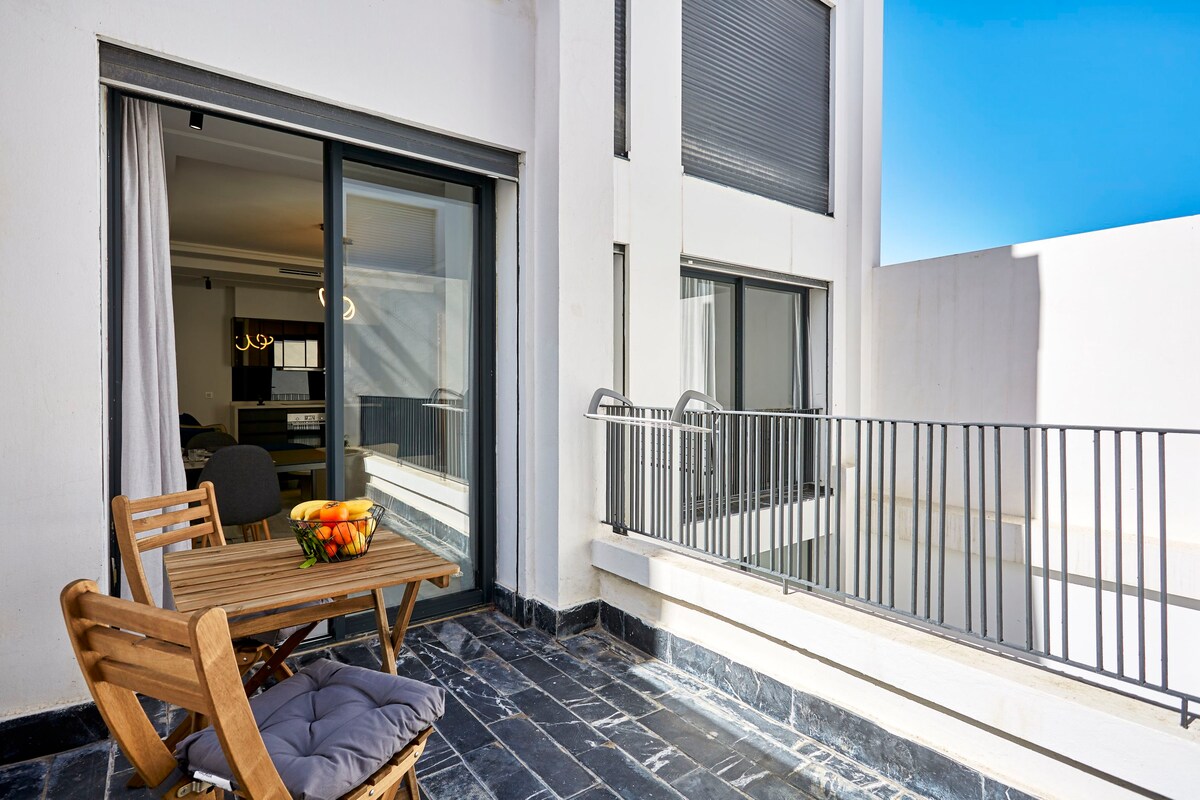 The terrace features a small wooden table with two cushioned chairs, complemented by a bowl of fresh fruit. Sliding glass doors provide access to the living area, while a balustrade offers a view of the surroundings under a clear blue sky.
