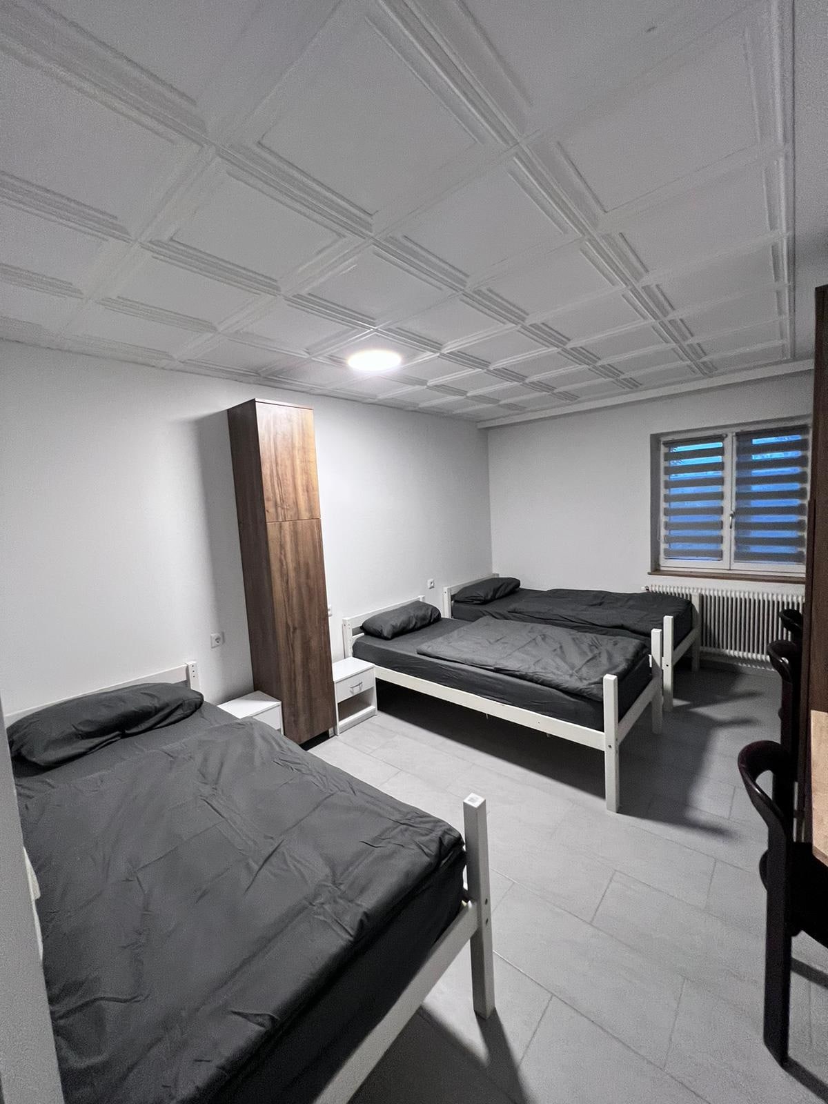 A well-lit guest room is equipped with three individual beds, each topped with dark bedding. A wooden partition is positioned in the center, and a window provides natural light, enhancing the spacious feel of the room. Simple decor contributes to a functional atmosphere.